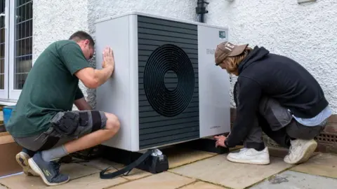 Getty Images Two men install a heat pump outside a house. Both are crouching either side of the heat pump. The man on the left is wearing a green t-shirt and shorts. The man on the right is wearing a black hoodie, trousers and a baseball cap.