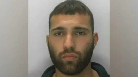 A police custody mugshot of Aaron Akram. He has short dark and facial hair. He is looking directly at the camera with a blank expression.