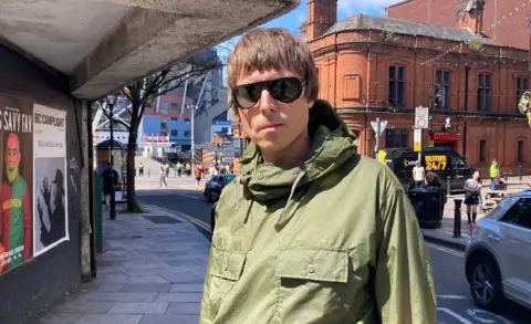 A picture of Glenn looking at the camera with a stern expression. He is wearing a khaki green parka jacket, black sunglasses and has short brown hair. He is standing in Cardiff town centre. Behind him is a red brick building and the Principality Stadium. 