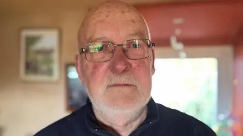 An older man with a bald head, white beard, moustache and glasses. He is wearing a navy zip-up sweatshirt and is looking at the camera with a serious expression. The background behind him is out of focus, but a large window and a painting hung on a wall to the right can just be seen.
