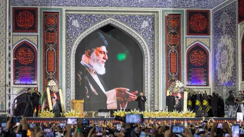 A crowd holds their phones up to take pictures at the funeral of Ayatollah Ali Khamenei, a picture of whom appears above a stage. 
