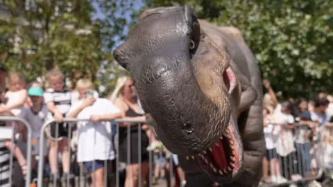 A brown t-rex puppet is looking at the camera. Its mouth is open and you can see its teeth. There are crowds of people behind a fence watching.