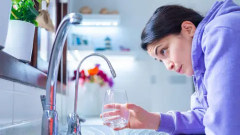 Getty Images Young woman in a purple hoodie leans over a kitchen sink, holding a glass under a mixer tap. She looks slightly concerned at the lack of water flow from the tap. There is another tap in the foreground.