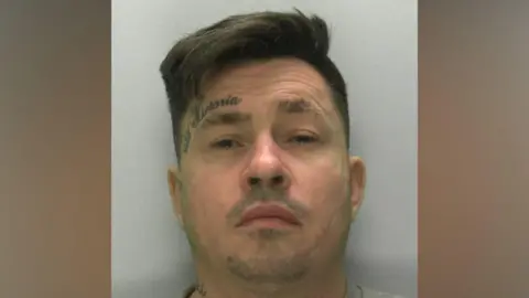 A mugshot of a man in his late 30s with quite a wrinkly forehead and a tattoo of a crown and the name "Victoria" above his left eye. He has brown hair swept over to one side. He is wearing a grey sweatshirt and standing in front of a light grey or white wall. He looks uncomfortable.