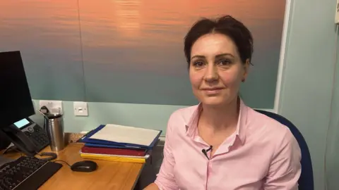 A woman with dark hair wearing a pink shirt sitting at a desk