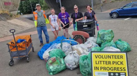 Peterborough Litter Wombles Five people - one male and four female are standing near an underpass. There are numerous bags of collected rubbish on the road in front of them and a yellow sign that says "volunteer litter pick in progress"