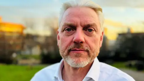 Man with grey beard and hair wearing white open-necked shirt, looking at camera, with grass, trees and buildings blurred in the background.