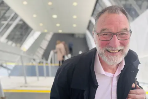 Colin Keillor, a man smiling at camera at top of stairs, he has a grey beard and glasses and is wearing a pink open-necked shirt on under a dark jacket.