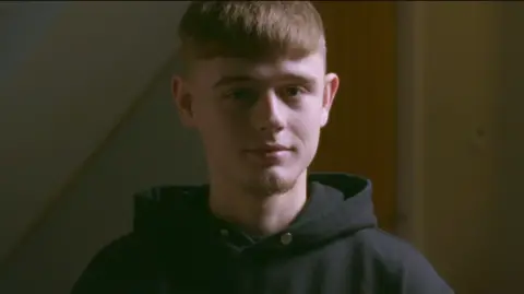 Cymru Wale stares a young man in the camera. It has short brown hair, very brief pieces on both sides. He has a distinct diming in the chin. He wears a dark convincing summit.