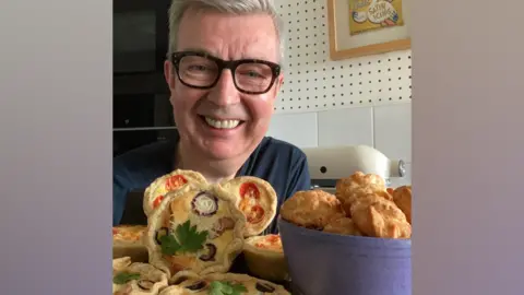 Howard Middleton Howard Middleton has short grey hair and is wearing brown framed glasses and a blue t-shirt. He is in a kitchen and is smiling at the camera with an array of baked good in front of him.