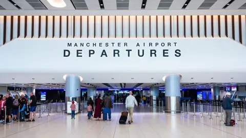 Manchester Airport departures hall, a large, white open space with a white tiled floor, people queuing to the left and other people either walking through or standing and waiting. A large sign reading Manchester Airport Departures is overhead.