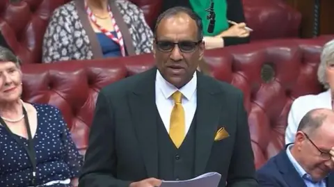 A man stands in a row of red leather seats. He is wearing glasses, a dark suit and yellow tie and is holding papers stapled together between his hands. The man next to him has his face in his hands while the two women in the row behind him look at him from either side.