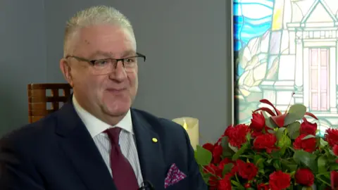 A grey haired man in a blue suit is sitting in front of a bunch of red roses and a lit candle. There is a stained-glass window in the background.