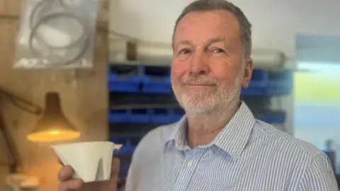 A man wearing a striped white and blue shirt with grey hair and a beard is smiling at the camera. He is holding a white container.
