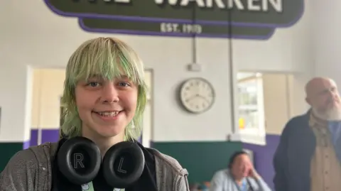 Cricket, who identifies as they/them, has green hair and has headphones around the neck.