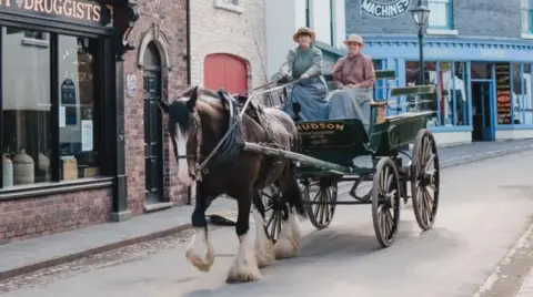 Ironbridge Gorge Museum Trust A black four-wheeled cart, pulled by a large dark horse and ridden by two women in Victorian clothing on a narrow street with a number of brick-walled shops behind it
