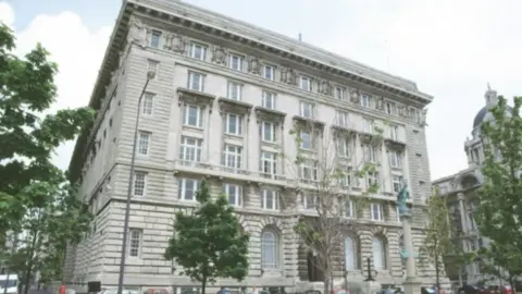 Image shows the Cunard Building, the headquarters of Liverpool City Council. It is an early 20th century Greco-Roman-style building of six storeys, with a Portland stone exterior. Relatively young trees are growing in the foreground.