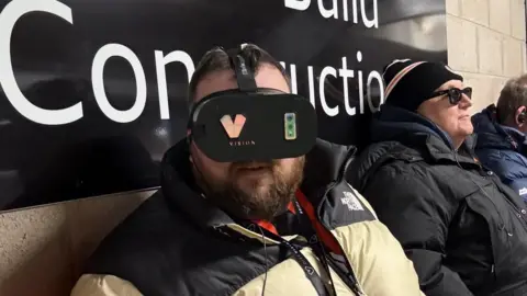 Jon Attenborough, a 36-year-old man with dark hair and a beard, wearing a puffer jacket and a black headset over his eyes. A fellow supporter wearing a jacket, hat and sunglasses is to his left.