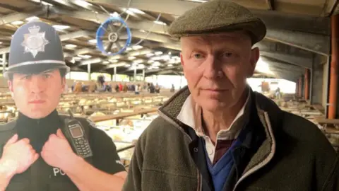A man in a brown flat cap and brown jacket in a large barn with animals in the background. There is a life-sized cut out of a police officer to his right hand side.