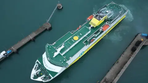 An aerial image of a DFDS ferry on the water. It has a largely green floor surface, and white and yellow sides. 