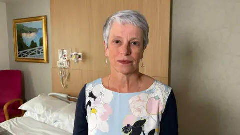 Ben Schofield/BBC Sharon Allen looking into camera in front of a hospice bed. She has short grey hair and is wearing earings. She is also wearing a light blue top with white, pink and black flowers on it and dark sleeves.
