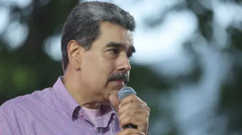 EPA Nicolás Maduro speaks into a microphone during the Great March of Communal Youth in support of his government in Caracas, Venezuela, 13 November 2025.
He is wearing a pink T-shirt and a pink-and-blue shirt and clutching the microphone with both hands. 