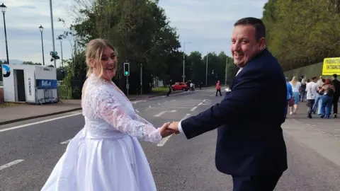 A woman, with blonde hair, tied back, wearing a white wedding dress. She is holding her husband's hand, who is wearing a black wedding suit. They are standing in the middle of a road. 