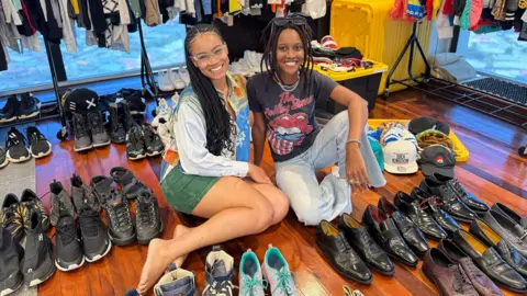 Atas perkenan Aliyah Clarke Aliyah Clarke dan Kaleen Sanois menjual pakaian Machel Montano di toko pop-up. Mereka duduk di lantai, dikelilingi oleh sepasang sepatu.

