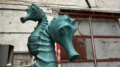 Andrew Turner/BBC A sculpture of a sea horse, which is a mottled weathered copper colour, standing in a warehouse, with dirty white walls and rusting steel doors behind it.