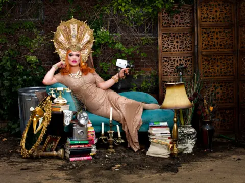 Magnus Hastings Uma drag queen com cabelos ruivos na altura dos ombros se pendura em uma espreguiçadeira enquanto usa um cocar dourado. Ela está cercada por artefatos antigos e segura uma velha câmera vintage