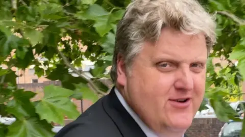 Gregory Hill, a man in a black suit jacket, white shirt and striped tie. He has blonde hair and is pictured outside court in Norwich.