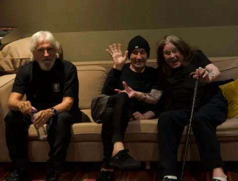 Ozzy Osbourne Three men sit on a beige sofa. They are all wearing all-black outfits. The man on the left has grey hair and a grey beard. The man in the centre has a black beanie hat. The man on the right has shoulder-length greying hair and a walking stick.