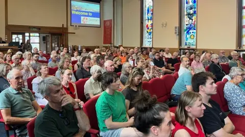 Andy Watson/BBC A modern church - with stained glass windows visible - is packed with volunteers of all different ages in its red chairs. They look with intensity and possibly mild concern towards the front of the room.
