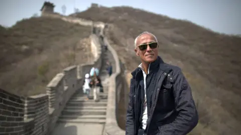 Craig Hastings A recent photo of Andrew Ridgeley, sitting against the Great Wall of China. He has grey cropped hair, is wearing sunglasses, a navy jacket and white top.