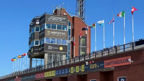 The tower at the Isle of Man TT grandstand. It is a red brick structure with large glass window all round it. There is a row of international flag on the front of the grandstand below it.