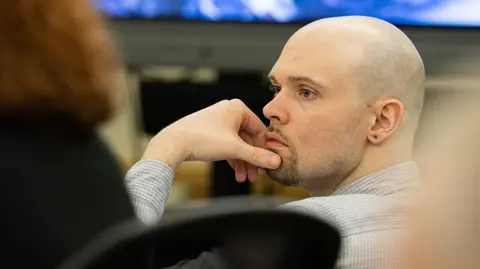 A bald man in a checked shirt looks into the distance with his left hand on his bearded chin. His left ear has a black stud earring