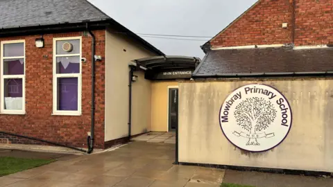 The main entrance of Mowbray Primary School has a large circular logo of a tree against a white background next to the main doors.