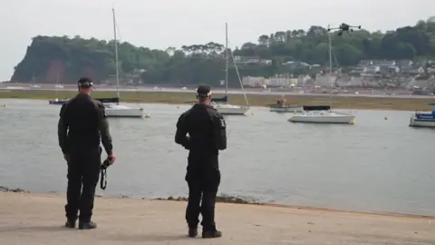 BBC Two Devon and Cornwall Police officers stood by a quayside operating a drone. There is some boats anchored up on the water and in the distance there are some cliffs and buildings. 