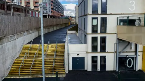Robin Stott/Geograph White building of several storeys with floor numbers on the glass of the stairwell. There are yellow exterior steps to the left, with metal handrails. There is a six-storey brick-built building in the background.