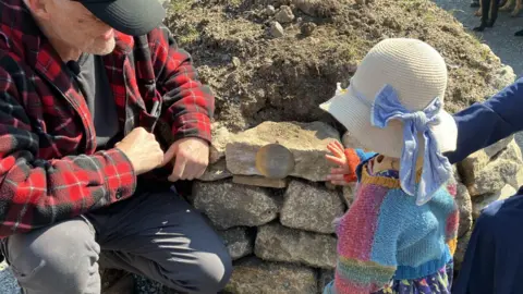 A man wearing a lumber jacket and crouched down leaning against a Cornishs hedge is looking at a young girl who is wearing a bonnet with a blue ribbon who is touching a piece of granite with a plaque on it. She is wearing a multi-coloured cardigan. 