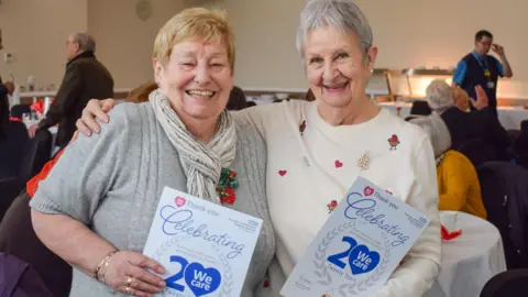 Two women of mature years stand smiling at the camera, with their arms round each other. One has short grey hair and wears a white jumper with hearts and robins on it. The other has blonde hair and wears a grey jumper. They both hold certificates which say "Celebrating 20 Years".