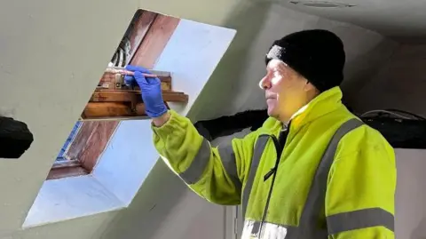 Ickleton Lionhearted A man is stood inside a room while painting a window fame. He wears a woolly hat, blue latex gloves and a thick yellow high visibility jacket. 