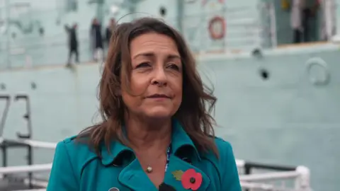 BBC Kent County Council leader Linden Kemkaran is interviewed by the BBC, wearing a turquoise overcoat with a memorial poppy in the lapel, standing in front of a boat at the Historic Dockyard in Chatham.