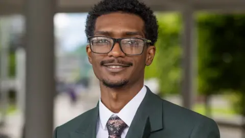 Northumbria University Ali Alamin is a young man in his 20s wearing a white shirt and tie and glasses. He has dark hair and a moustache and a small beard.