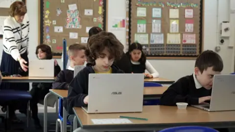BBC School children sitting at desks in a classroom setting with laptops.