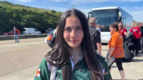 Alexia Nascimento is standing in front of a coach smiling into the camera. She is wearing Guernsey kit.