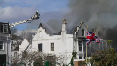 Black smoke coming from the derelict hotel in Torquay. The smoke is black and is filling the sky. A firefighter is on an ariel ladder to the right fighting the fire.