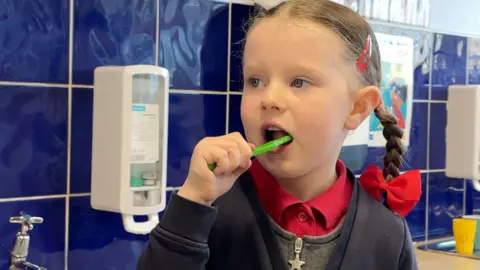 A five-year-old girl with her hair in plaits with red bows is standing in front of a sink, cleaning her teeth with a green toothbrush