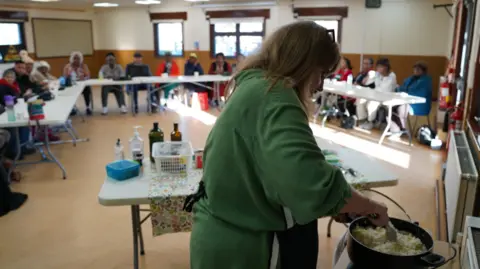 A woman in a green shirt is over a black pot stiring rice. She is in a community hall that has three long tables with people sat around it.