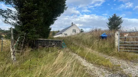 The house is at the end of a track. There is a wire fence and a large tree on the left, and a metal gate and smaller tree on opposite side of the track.
